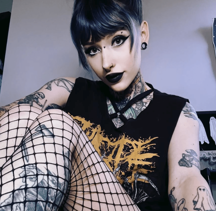 Goth OnlyFans model Gothmomlilli in a band tee and fishnets with black lipstick and tattoo sleeves